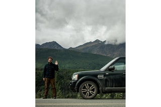 A man waving at the camera with a car on the left of shot and mountain scenery in the background.