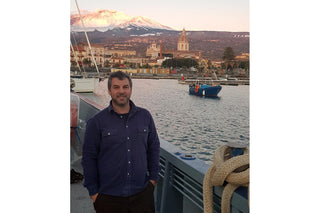 A man standing on a boat with a port city and mountains in the background.