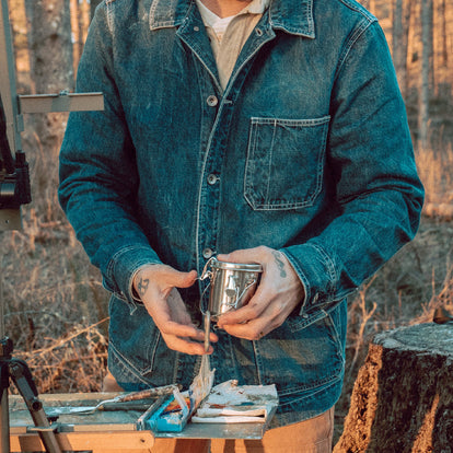 Zach standing amongst easels in his studio while wearing The Moor Sweater in Washed Indigo.