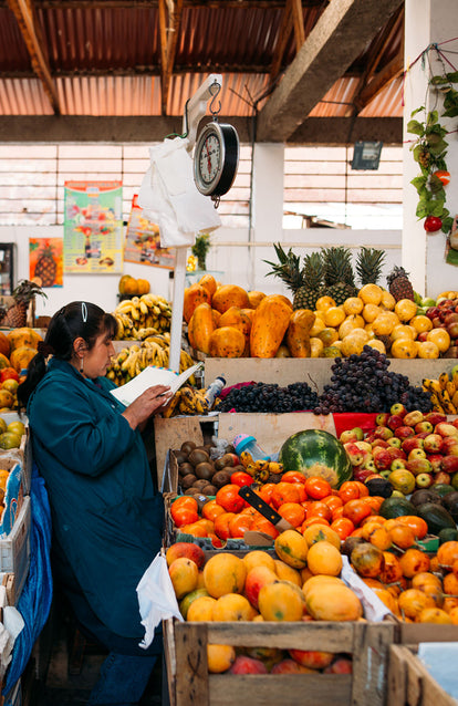 A woman checking a ledger, standing in a packed tropical fruit stand.