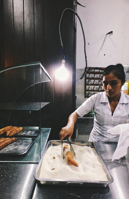 A white-coated baker, using tongs to dip pastry in a pan full of white sugar.