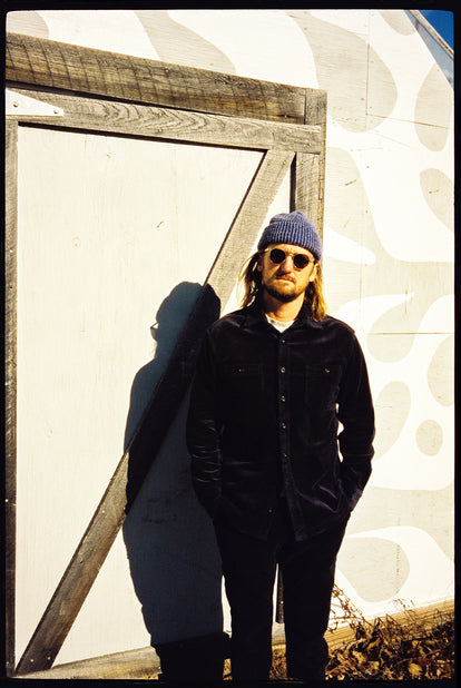 Long-haired guy dressed in dark shirt, dark pants, dark shades, against a white and wooden-beamed wall.