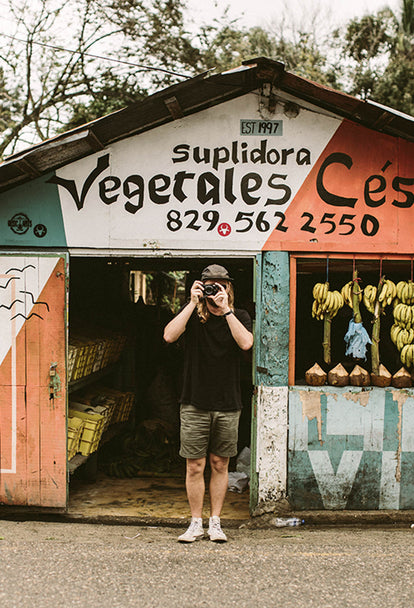 A photogrpaher stadning outside a rural fruit stand, colorfully painted with bunches of bananas hanging.