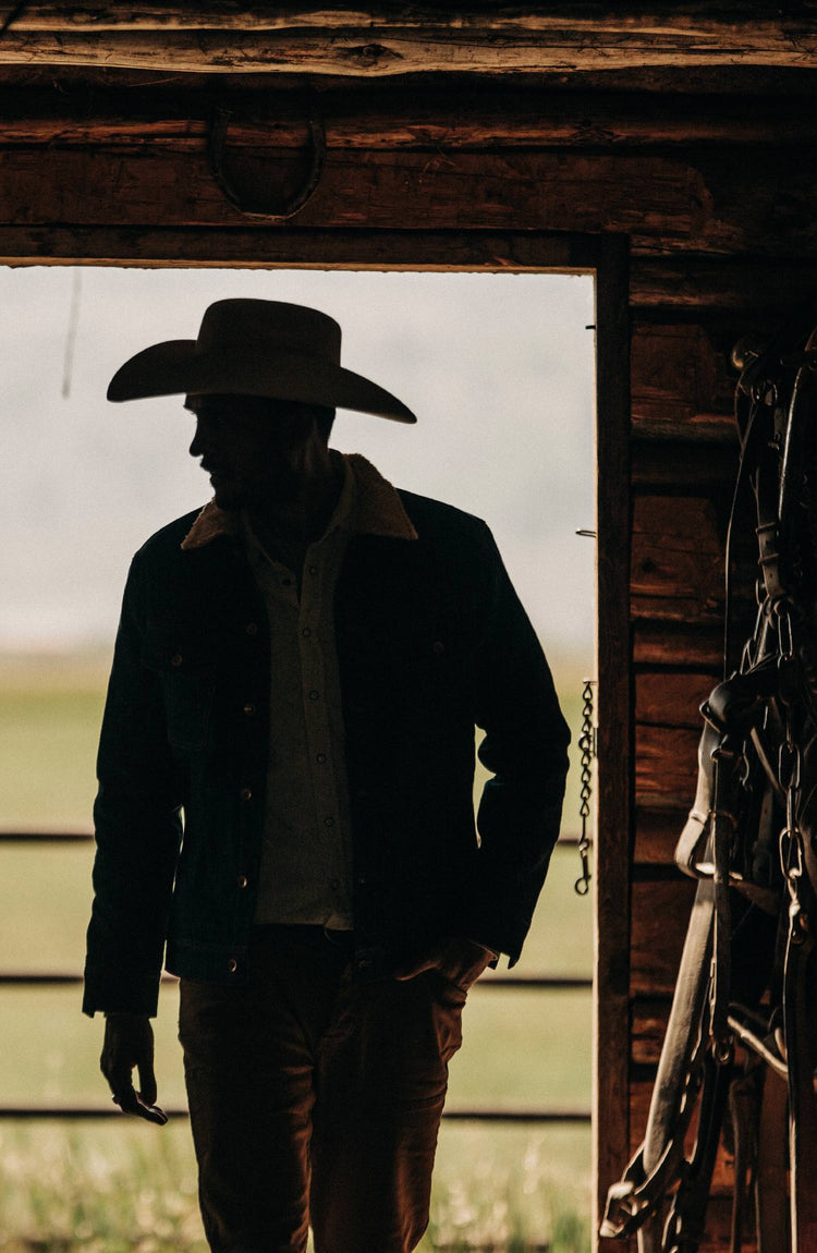 Model wearing The Lined Long Haul Jacket in Green Cast Denim in a stable with saddles hanging on the opposing walls