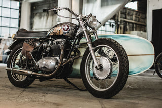 A highly customized vintage motorbike with a surfboard mounted in a carrier on the far side.