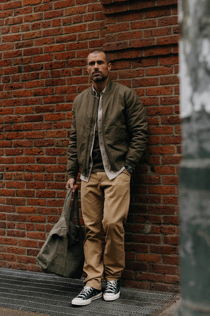 Model standing against a brick wall in The Bomber Jacket in Fatigue Olive and the Foundation Pant in Khaki