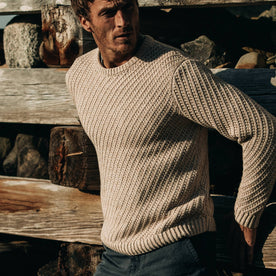 fit model wearing The Adirondack Sweater in Natural, leaning against fence, Knits by Taylor Stitch