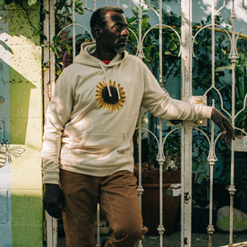 fit model leaning against fence wearing The Fillmore Hoodie in Sunflower Embroidery, Knits by Taylor Stitch