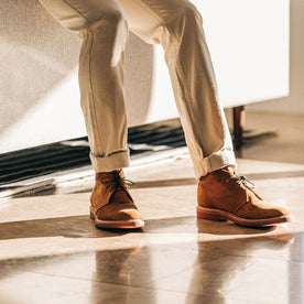 The Unlined Chukka in Butterscotch Weatherproof Suede—pants cuffed, sitting against couch, Footwear by Taylor Stitch